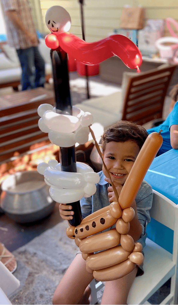A young child sits on a chair, smiling and holding a large pirate ship balloon sculpture crafted by a talented balloon artist, complete with a red flag and white skull and crossbones sail.