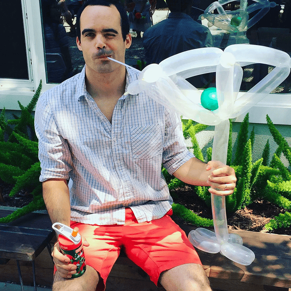 A man in a striped shirt and red shorts sits on a bench in Los Angeles, holding a large cocktail-shaped balloon and sipping through a straw, with lush greenery in the background.