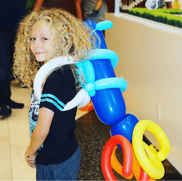 Child with curly blond hair wears a black t-shirt and a rocket backpack made from colorful balloons, standing indoors with vibrant balloon decor and smiling at the camera.