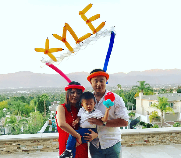A family of three poses on a balcony with mountain views in Los Angeles; the adults wear balloon hats, and a festive balloon sign spelling "XYLE" is held above them, showcasing creative balloon installations.