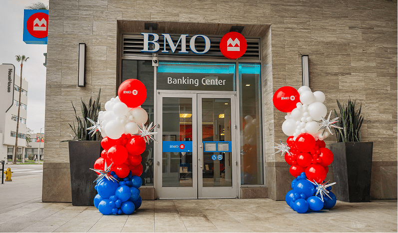 Entrance of a BMO banking center featuring stunning balloon decor, with red, white, and blue balloons artfully arranged on either side of the glass doors.