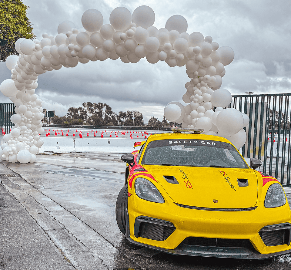 A yellow safety car is parked on a wet track near a stunning balloon arch made of white balloons, with traffic cones and a cloudy sky in the background.