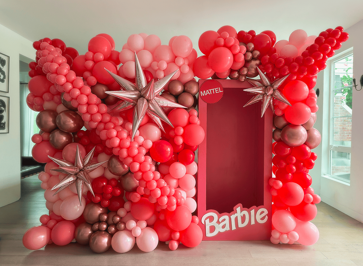 A life-sized Barbie doll box display is surrounded by clusters of pink, red, and metallic balloon decor, with large silver star balloons accenting this stunning balloon installation.