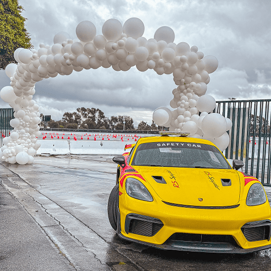 A yellow safety car is parked on a wet street near a Los Angeles racetrack entrance, decorated with an arch of white balloons under a cloudy sky.