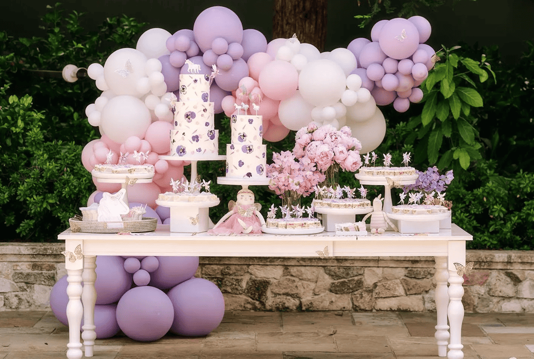 A decorated dessert table with pastel purple and pink cakes, sweets, and flowers, set outdoors in Los Angeles with a backdrop of matching balloon decor installations and lush greenery.