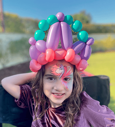 Young child with long hair wears a colorful balloon crown, crafted by a talented balloon artist, and has a painted unicorn on their face, standing outdoors with a blurred background.