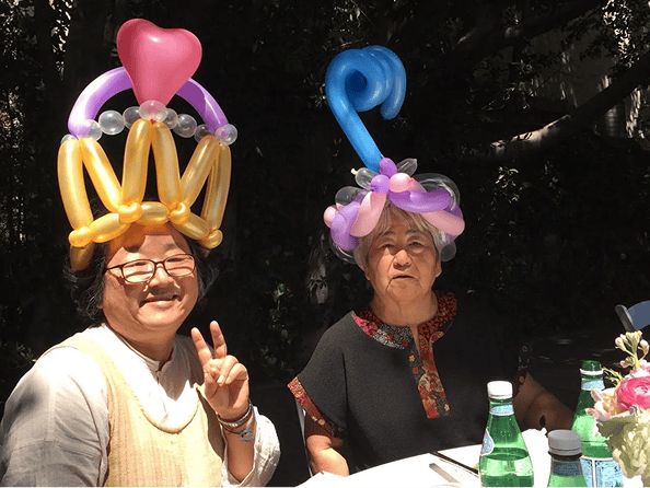 Two women sit at a table outdoors in Los Angeles, each wearing colorful balloon hats; one smiles and flashes a peace sign while the other looks on with a neutral expression. Bottled water sits amid their playful balloon installations.