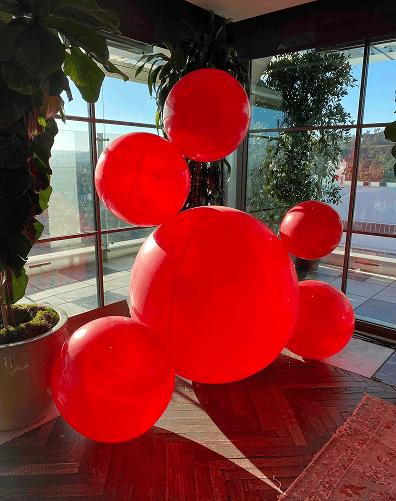 Five large red balloons of varying sizes are clustered together indoors near large windows with sunlight streaming in, showcasing the creativity of a balloon artist.