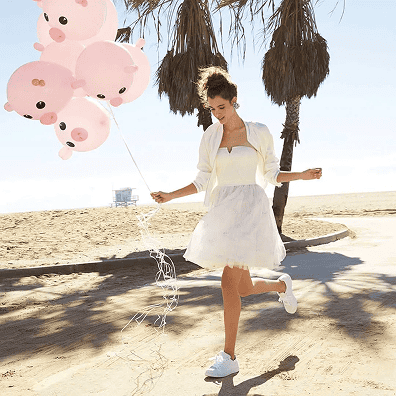 Woman in a white dress and sneakers holds a bunch of pink pig-shaped balloons crafted by a talented balloon artist while walking on a sandy path near palm trees.