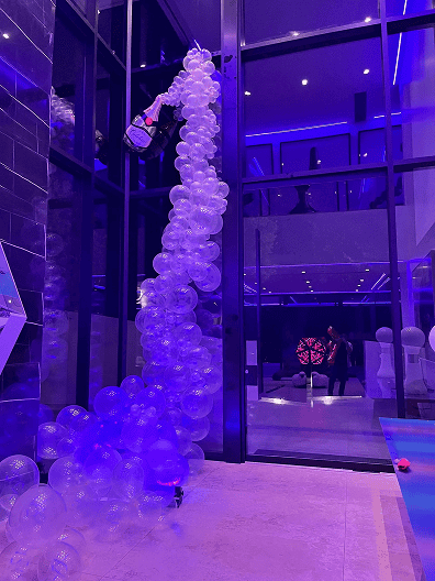 A tall column of transparent balloons rises from floor to ceiling in a modern, glass-walled room with blue-purple lighting—an impressive balloon installation perfect for a corporate event. Two people are visible in the background.