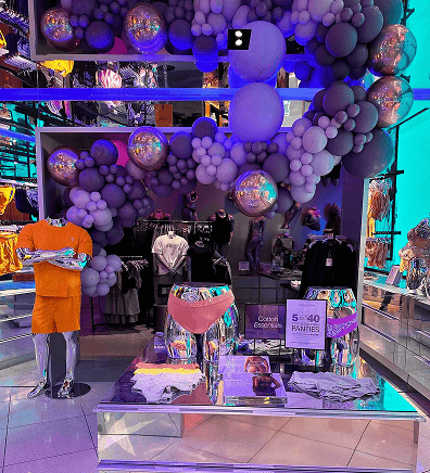 Clothing store display with metallic mannequins, pink and purple underwear, folded clothes, and stunning balloon decor by a skilled balloon artist overhead.