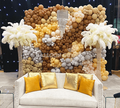 A white sofa with gold pillows sits before a stunning balloon installation in brown, gold, and cream tones, flanked by two tall feather centerpieces and a chandelier overhead—perfect for a chic Los Angeles corporate event.