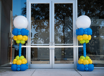Two glass doors are decorated for a corporate event with a white ribbon and two vibrant balloon installations in blue, yellow, and green, each topped with a large white balloon.