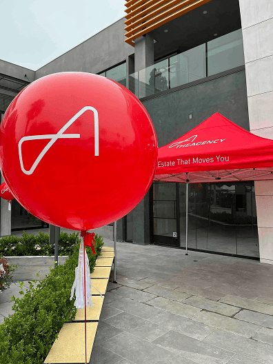A large red balloon with a white stylized "A" and a matching red tent with "Estate That Moves You" text set the scene for a vibrant corporate event outside a modern building.