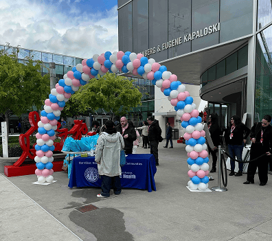People gather at an outdoor event under vibrant balloon decor, featuring a pink, blue, and white arch near a table with a blue cloth labeled “Department of Health.” A building is in the background.