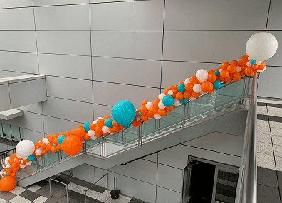 A glass stair railing is decorated with clusters of orange, white, and blue balloon decor for a corporate event in a modern indoor Los Angeles setting.
