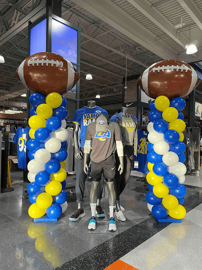 Two balloon installations with football toppers flank a mannequin dressed in LA Rams gear, adding vibrant balloon decor to a Los Angeles retail store filled with Rams merchandise.