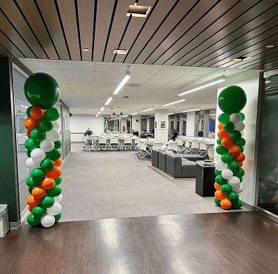 Two columns of green, orange, and white balloon installations stand at the entrance of a modern, open office space with tables, chairs, and lounge seating.