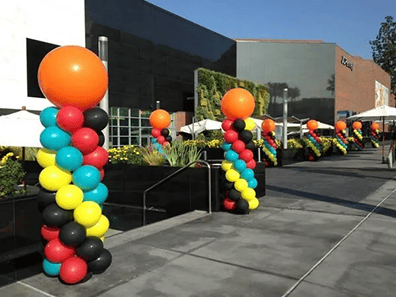 Colorful balloon columns crafted by a skilled balloon artist, featuring orange tops and vibrant red, yellow, black, and blue balloons, line an outdoor walkway beside a modern building on a sunny day.