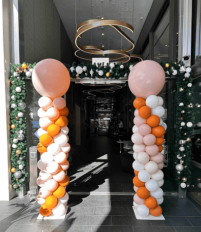 Entrance decorated with two balloon columns in peach, orange, and white, plus a garland with ornaments overhead—perfect for a corporate event. Modern lighting fixtures visible inside highlight the impressive balloon installations.