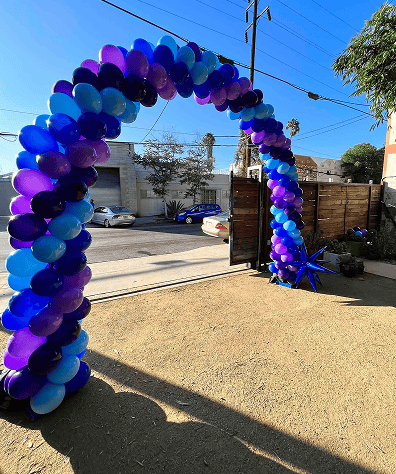 A large arch made of blue and purple balloons, perfect for a Los Angeles corporate event, stands in an outdoor yard near an open gate, with cars and buildings visible in the background.