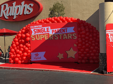 A red balloon arch and backdrop with festive balloon decor outside Ralphs display the message "Celebrating Single Parent Superstars," featuring Yoplait branding and gold stars.