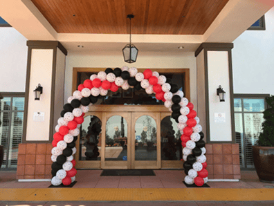 A large arch made of red, white, and black balloons stands at the entrance of a building with double wooden doors and a covered entryway, showcasing stunning balloon decor perfect for events in Los Angeles.