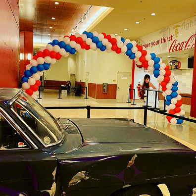 A damaged vintage car is displayed indoors beside a striking red, white, and blue balloon decor arch, with black stanchions and concession stands in the background.