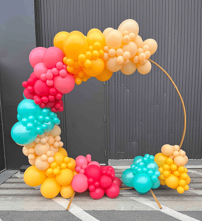 A large circular balloon arch featuring vibrant clusters of yellow, red, orange, and teal balloons creates stunning balloon decor against a gray corrugated wall outdoors in Los Angeles.