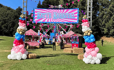 Outdoor carnival setup with a large sign reading "Katy's Karnival," striped tents, colorful balloon installations, hay bales, and people standing on grass.