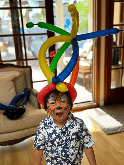 A young child with face paint resembling a tiger wears a colorful balloon hat and a floral shirt, standing indoors in Los Angeles amid vibrant balloon decor, in front of a sofa and large windows.