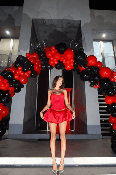 A woman in a short red dress stands under a stunning balloon decor arch of red and black balloons in front of a modern building at night.