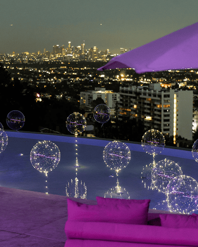 A pool with floating illuminated spheres overlooks the Los Angeles skyline at dusk, with purple lounge seating and an umbrella in the foreground, evoking the feel of elegant balloon installations.