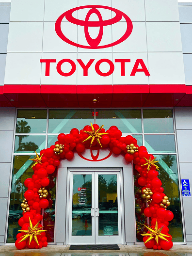 Entrance of a Toyota dealership decorated with stunning balloon decor, featuring a red and gold balloon arch and large gold star ornaments.