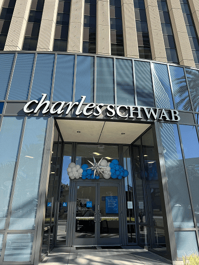 Front entrance of a Charles Schwab office building in Los Angeles with glass doors, the company name above, and festive blue and white balloon decor displayed over the entryway.