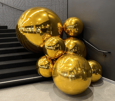 A cluster of large, shiny gold spheres creates stunning balloon decor at the base of a modern staircase with dark steps and gray walls, perfect for stylish interiors in Los Angeles.