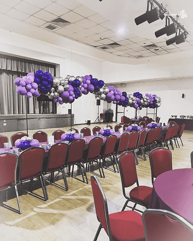 Rows of red chairs and tables with purple tablecloths are arranged in a hall, beautifully decorated with purple and silver balloons by a talented balloon artist in Los Angeles.
