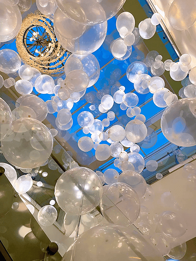 Large clear balloons are suspended in the air inside a building during a corporate event in Los Angeles, with a chandelier above and a blue sky visible through a skylight.