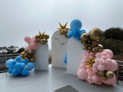 Decorative balloon arch display with pink, blue, and gold balloons, featuring two white panels and a "Boy or Girl?" sign, set outdoors on a rooftop—perfect for stylish balloon installations at your next celebration.
