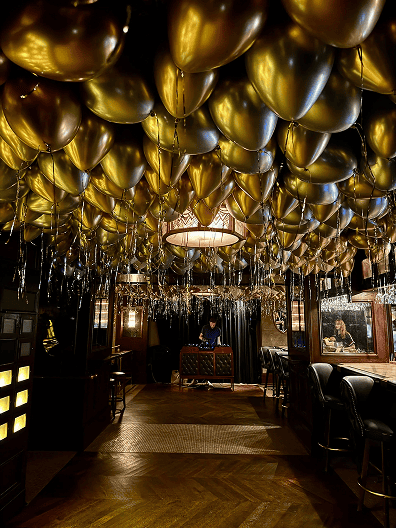 A bar with a wooden floor and black leather stools features stunning balloon installations, with gold balloons and hanging ribbons covering the ceiling. A person stands behind a counter in the background.