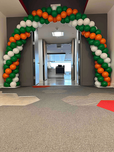 An archway of orange, white, and green balloon decor stands at the entrance of an office hallway, with open doors leading to a desk in the background.