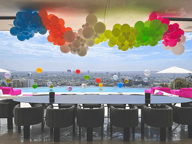 A modern outdoor dining area in Los Angeles overlooks a cityscape, with colorful balloon installations decorating the ceiling and floating by the poolside.