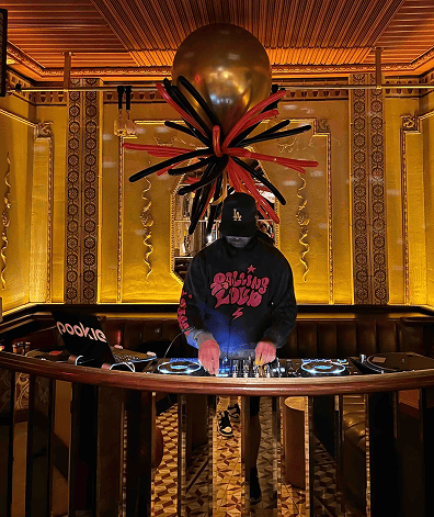 A DJ wearing a mask and cap performs at a small booth in an ornate room with golden walls, surrounded by vibrant balloon decor and a creative balloon installation overhead.