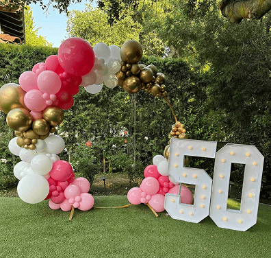 A balloon artist in Los Angeles created a stunning pink, white, and gold balloon arch next to large light-up numbers "50" on a lawn, surrounded by trees.