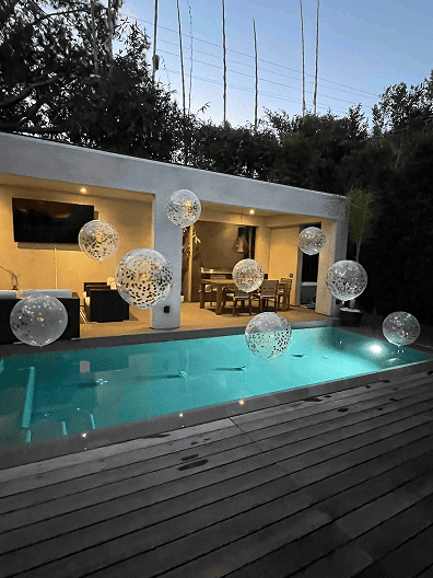A modern backyard with a small illuminated pool, floating decorative spheres resembling balloon decor, a wooden deck, and an open dining area inside a white structure.