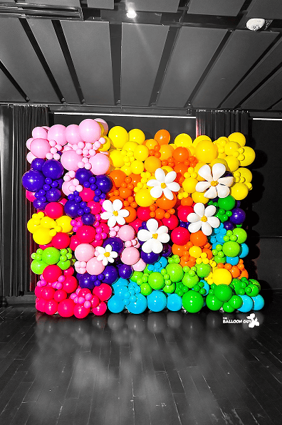 A colorful wall of balloon decor arranged in a rainbow pattern with white and yellow flower-shaped balloons, set on a dark stage—perfect for eye-catching balloon installations in Los Angeles.