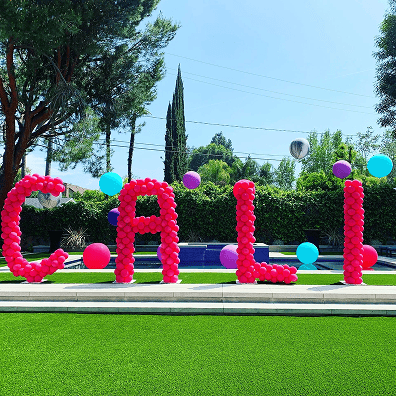 Large balloon letters spelling "CALI" create striking balloon decor on grass by a pool, surrounded by trees, blue sky, and additional balloons in the background.