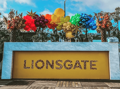 A marble counter with a bright yellow panel displaying the word "LIONSGATE," decorated with colorful balloons and star ornaments in the background, perfect for a festive corporate event or featuring a talented balloon artist.