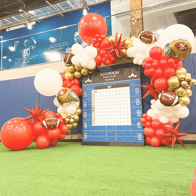 A Los Angeles-inspired football-themed balloon arch, featuring red, gold, and white balloons with football-shaped details and a scoreboard display, creates a striking balloon installation on artificial turf indoors.