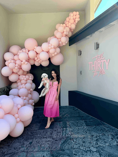 Woman in a pink dress holding a small white dog stands next to a large pink balloon arch, created by a talented balloon artist in Los Angeles, and a sign that reads "Talk Thirty to Me.